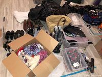 Wide view of lot showing shoes lined along wall, cardboard box with folded clothing, purses, plastic storage bins containing clothes