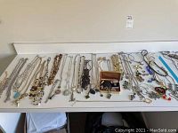 Wide shot showing the full assortment of jewelry laid out on a white table, including necklaces, pins, bracelets with mixed materials.