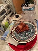 Photo of dishware including two patterned ceramic platters with floral designs, wooden salad utensils, a ceramic covered dish, and a clear plastic drink server with kitchen items in background.