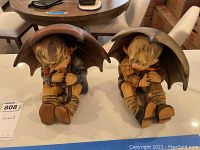 Two 8-inch Hummel boy figurines sitting with umbrellas held above them, viewed from the front.