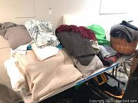 Assorted linens, blankets, comforters, pillows, and a wicker basket piled on a table or surface.