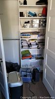 View of shelving with various office supplies including notebooks, file folders, storage bins, paper supplies, shredder and electronics on floor.