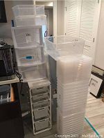 Overall view of tall stacks of clear plastic storage bins and a white storage bin rack with drawers in indoor setting.