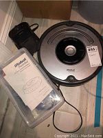 Photo of the Roomba 655 robot vacuum, charging dock, and user manual in a plastic container.