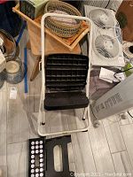 Folding step stool with white metal frame and black textured plastic steps with woven baskets and trays on a small wooden table behind it.