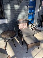 Photo of the outdoor wicker chair with cushions, ottoman, and round outdoor table on concrete patio against brick wall and blue door.