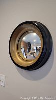 Photo of round convex wall mirror, showing the black exterior frame and metallic gold interior rim around convex glass, hung on beige wall near light switch.
