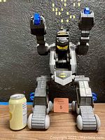 Front view of the Batbot robot next to a soda can for size reference, showing gray and blue plastic body with Batman logo on chest, robot stands about 19 inches tall.