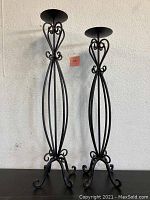 Front view of two black metal tall candle stick holders with scroll details on elliptical frames and curled feet.