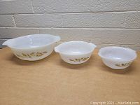 Three vintage white glass mixing bowls of graduated sizes featuring golden wheat patterns shown on a wood surface with a brick background.