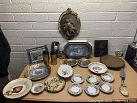 Table display of assorted vintage items including decorative plates with floral and bird designs, framed art prints, metal tray, brass tumblers, and an oval wall plaque with classical figure in relief.