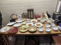 Wide view of assorted ceramic plates, bowls, cups, and decorative figurines on a wooden table against a white brick wall.