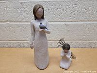 Photo of a standing Willow Tree figurine holding a bird and a smaller kneeling angel figurine with wire wings on a beige floor against a white textured wall.