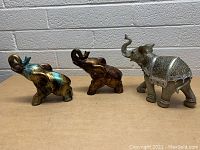 Photo showing three elephant figurines: one silver-tone with detailed patterns, two brass-tone in abstract style.
