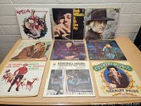 Front view of nine assorted country music vinyl record covers arranged in three rows on a wooden table against a white brick background.
