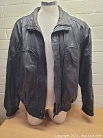 Front view of men's black leather jacket on mannequin, shows zipper, snap buttons, pockets, and slight white marks