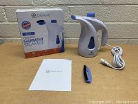 Deneve Blue Portable Garment Steamer shown with original box, user manual, fabric brush attachment, and power cord laid out on a table in front of a brick wall.
