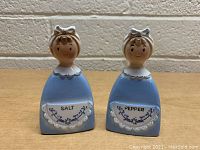 Front view of salt and pepper shakers, showing blue dresses, aprons with 'SALT' and 'PEPPER' text and floral design, and smiling female faces with headscarves.