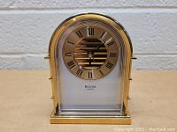 Front view of the Bulova gilt mantle clock showing circular face with Roman numerals and center stripe design under glass.