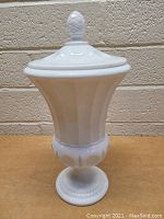 Front view of a vintage white opaque milk glass dish with lid and pineapple finial on top, placed against a brick wall background.