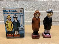 Front view of the vintage nautical-themed salt and pepper shakers standing beside their original box which features matching artwork.