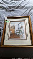 Framed limited edition print showing an interior scene with wicker chair, wood stove, and cupboard. Signed and numbered with edition number 308/390 and artist's signature visible.