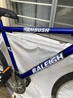Close-up of the blue bike frame showing 'Ambush' and 'Raleigh' decals with some wear.
