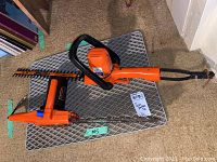 Black & Decker electric hedge trimmer with orange body and 16-inch blade on a wire rack indoors.
