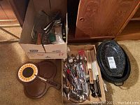 Two boxes containing assorted flatware, a sunflower design cheese board, and a black speckled roasting pan