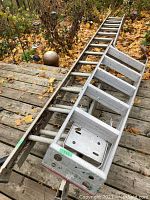 Two aluminum ladders lying on a wooden deck covered with autumn leaves, showing an extension ladder and a 6-foot step ladder with a platform on top.