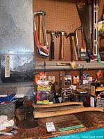 View of axes, saws, hammers hung on pegboard with various tool boxes and hardware supplies on table below