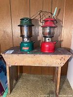 Photo showing two vintage Coleman lanterns, one green and one red, placed on top of a worn wooden bench against wood-paneled wall.