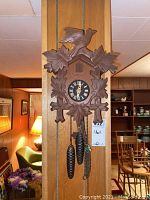 Full view of the vintage wooden cuckoo clock mounted on a wood-paneled wall, showing the carved bird atop the clock and pinecone weights hanging below.