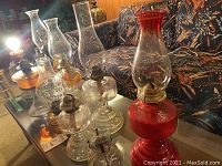 Overview of 9 vintage glass oil lamps arranged on a glass table, showcasing different sizes, colors, shapes, and chimney styles.