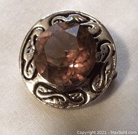 Front view of round silver tone brooch with central orange faceted stone and engraved swirling design.