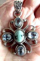 Front view of the pendant showing the turquoise center stone surrounded by four blue topaz stones and open work sterling silver design with ornate bail held in hand.