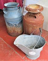 Blue milk can with eagle and star design showing rust, next to rusty milk can with lid and metal ash bucket in front.