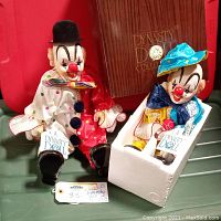 Two 1983 Dynasty Doll Collection sitting clown dolls, one in white polka dot suit with black bowler hat and one in yellow outfit with blue hat, positioned front of brown DYNASTY DOLL COLLECTION box.