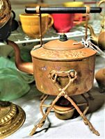 Copper antique teapot with warmer stand including black wooden handle and floral engraving on teapot body and lid. Warmer has three-legged scissor-style stand with burner below.