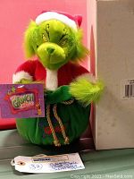 Green Grinch plush bear with Santa hat and jacket, holding a green bag labeled 'Grinch', new in box condition.