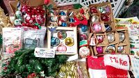 Overview photo showing multiple boxed glass ornaments, bows, stockings, artificial greenery, and small packaged decorations on a table.