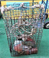 Green metal lobster trap with attached thick rope on top, red brick and orange and white buoy inside, photographed from front on green surface.