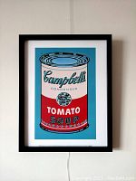 Framed lithograph hung on wall showing full image of Campbell's Tomato Soup can with black frame and white matting.
