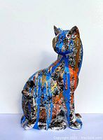 Front and side views of the sitting cat sculpture showing detailed blue, orange, gold, white, and black paint drips and splatters on a textured resin body.