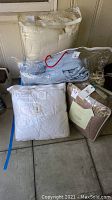 Several bedding items stacked including a white comforter, light blue bedding, and packaged King size Nostalgia quilt