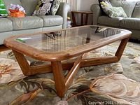 Angle view of the coffee table showing its solid wood base and glass top with rounded edges on a floral rug under living room sofas.