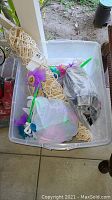Plastic bin containing a beige woven rope hammock, handheld windmills with purple flower blades and smiley face, miscellaneous items in plastic bags, and a grey item wrapped in plastic.