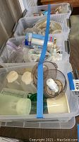 Photo of plastic bins filled with assorted kitchenware including white measuring spoons, cups, and food processor attachments secured with blue tape.