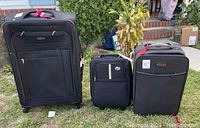 Photo of three soft suitcases: one large and two smaller carry-on size, all black with wheels and various zippered compartments, set outside on grass.
