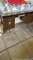Front view of wooden office desk showing the three drawers on the left and right side, black top surface with a 'Just Do It USA' sticker, and tiled floor beneath.